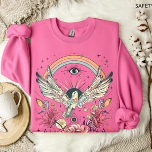 May include: A pink sweatshirt with a colorful graphic design featuring a bird with outstretched wings, a rainbow, an eye, and floral elements. The design is centered on the front of the sweatshirt.