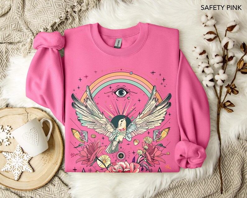 May include: A pink sweatshirt with a colorful graphic design featuring a bird with outstretched wings, a rainbow, an eye, and floral elements. The design is centered on the front of the sweatshirt.