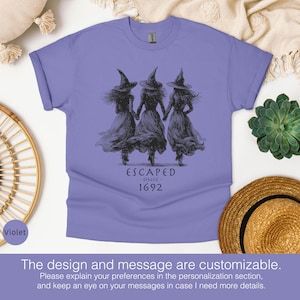 May include: A violet short-sleeved t-shirt featuring a black graphic of three figures wearing pointed hats and long dresses, holding hands. Below the figures, the text reads ESCAPED SINCE 1692 in a dark, stylized font.