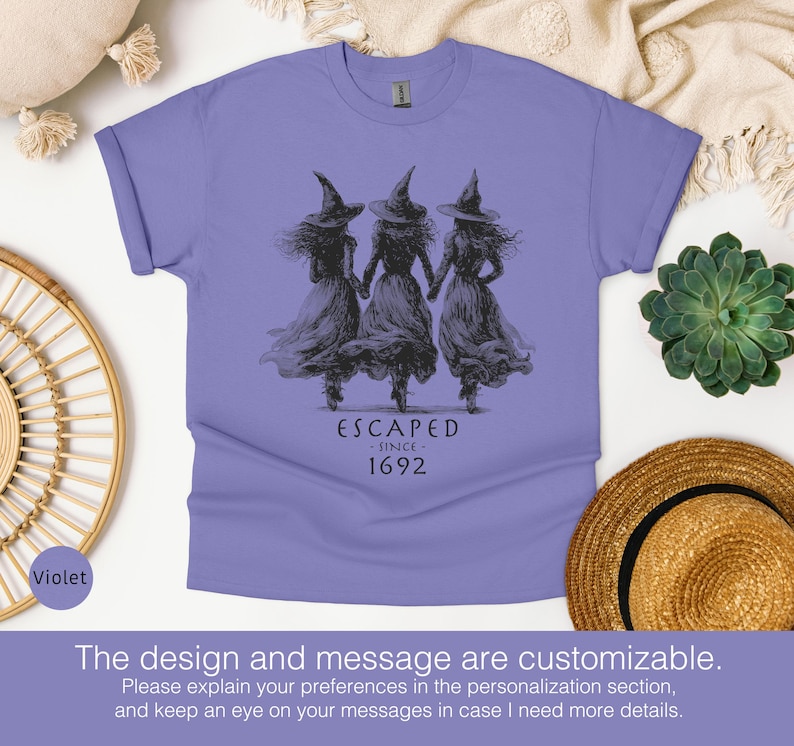 May include: A violet short-sleeved t-shirt featuring a black graphic of three figures wearing pointed hats and long dresses, holding hands. Below the figures, the text reads ESCAPED SINCE 1692 in a dark, stylized font.