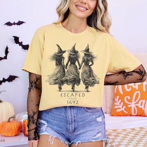 May include: A yellow haze t-shirt featuring a black graphic of three witches in pointed hats holding hands. Below the figures, the text reads ESCAPED SINCE 1692. The shirt is worn over a black long-sleeved mesh top with a floral pattern.
