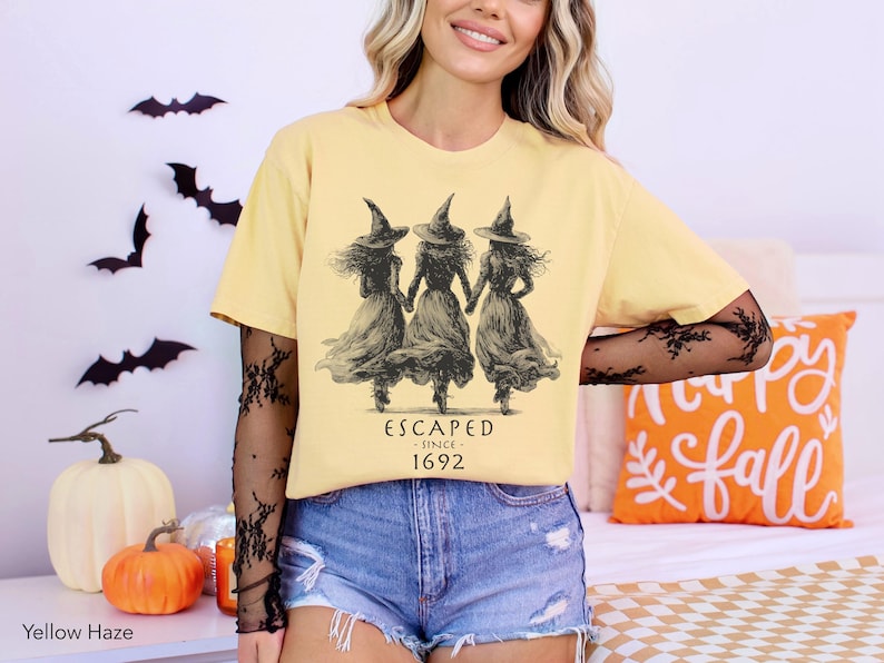 May include: A yellow haze t-shirt featuring a black graphic of three witches in pointed hats holding hands. Below the figures, the text reads ESCAPED SINCE 1692. The shirt is worn over a black long-sleeved mesh top with a floral pattern.