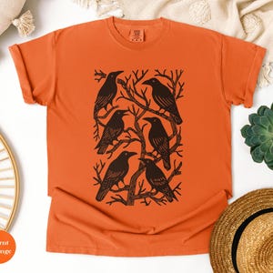 May include: Burnt orange t-shirt featuring a black graphic of six crows perched on tree branches. The crows are arranged in a rectangular pattern. The shirt is made of a soft material and has a classic crew neck.