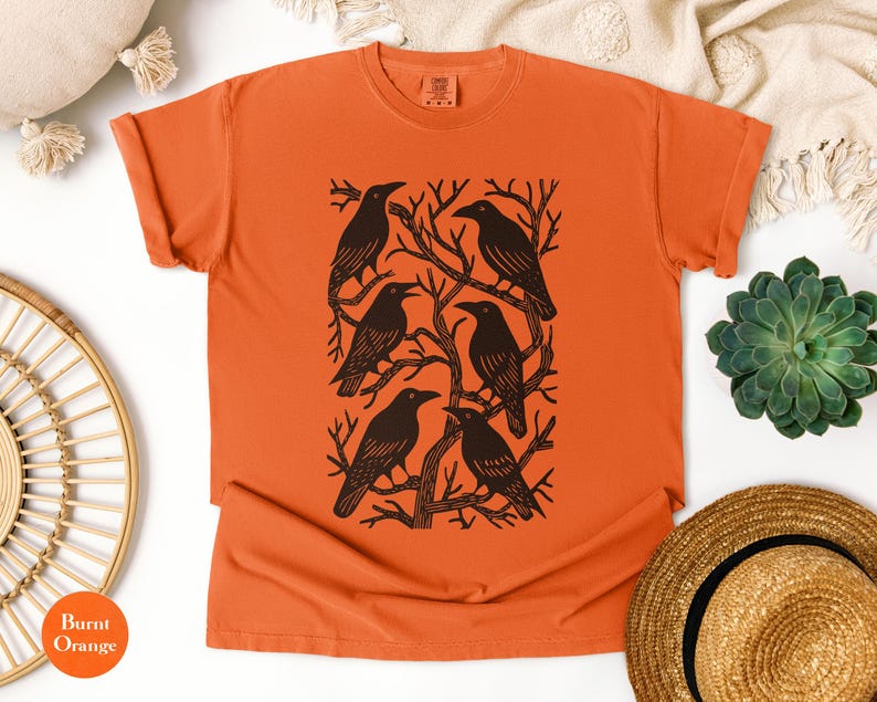 May include: Burnt orange t-shirt featuring a black graphic of six crows perched on tree branches. The crows are arranged in a rectangular pattern. The shirt is made of a soft material and has a classic crew neck.