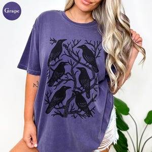 May include: A grape-colored t-shirt featuring a graphic of six black crows perched on bare tree branches. The crows are arranged in a circular pattern. The shirt has short sleeves and a crew neck.