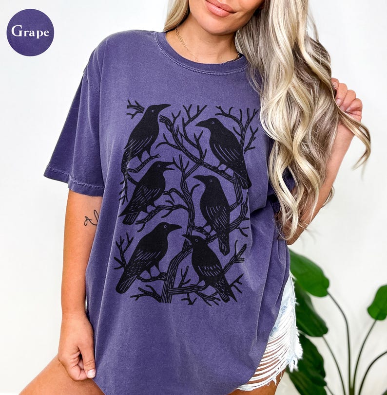 May include: A grape-colored t-shirt featuring a graphic of six black crows perched on bare tree branches. The crows are arranged in a circular pattern. The shirt has short sleeves and a crew neck.