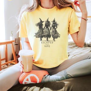May include: Butter-colored t-shirt featuring a graphic of three witches holding hands, with the text "ESCAPED SINCE 1692" in a vintage style. The shirt is a soft, comfortable material.