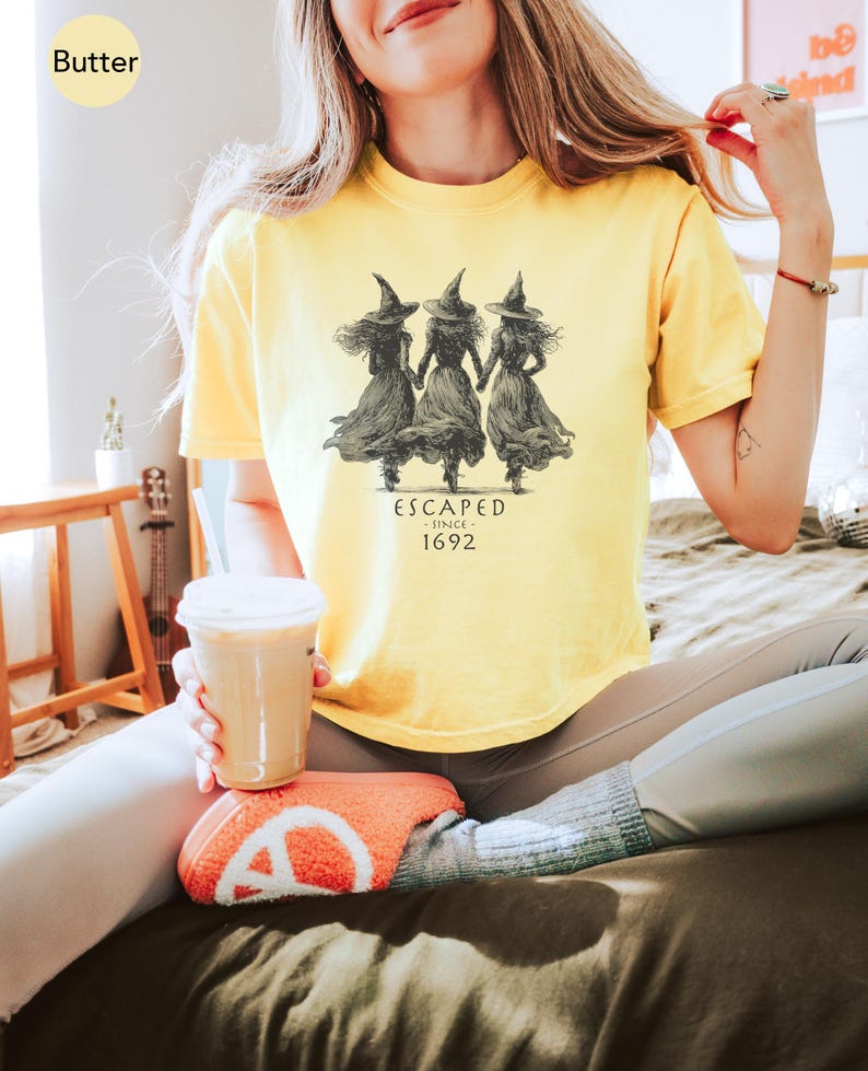 May include: Butter-colored t-shirt featuring a graphic of three witches holding hands, with the text "ESCAPED SINCE 1692" in a vintage style. The shirt is a soft, comfortable material.