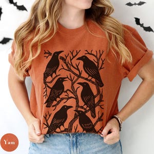 May include: An orange t-shirt featuring a graphic of six black crows perched on bare tree branches. The design is centered on the shirt, with the crows arranged in a circular pattern. The shirt has short sleeves and a relaxed fit.