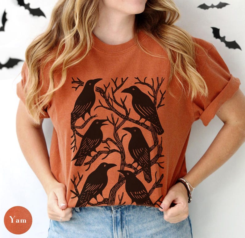 May include: An orange t-shirt featuring a graphic of six black crows perched on bare tree branches. The design is centered on the shirt, with the crows arranged in a circular pattern. The shirt has short sleeves and a relaxed fit.