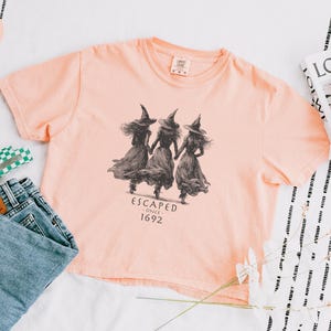 May include: A peach-colored t-shirt featuring a graphic of three witches walking together, with the text "ESCAPED SINCE 1692". The shirt is paired with blue jeans, a green and white hair clip, and a book.