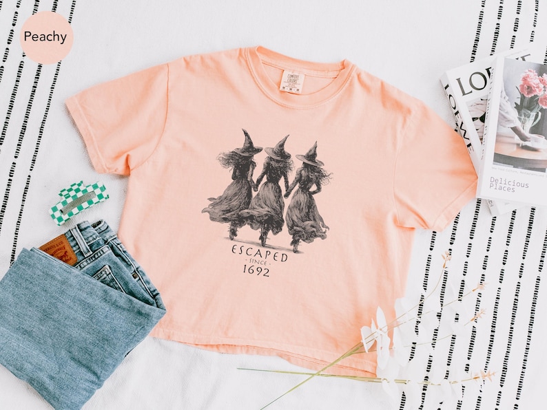 May include: A peach-colored t-shirt featuring a graphic of three witches walking together, with the text "ESCAPED SINCE 1692". The shirt is paired with blue jeans, a green and white hair clip, and a book.