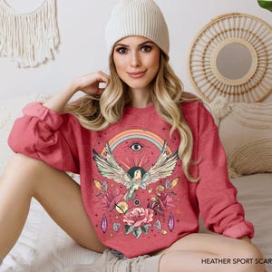 May include: A red sweatshirt with a colorful graphic print of a bird with an eye and rainbow. The text "HEATHER SPORT SCARLET RED" is printed on the bottom of the sweatshirt.