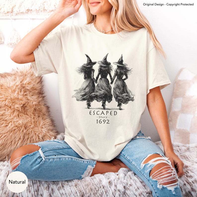 May include: A cream-colored short-sleeve t-shirt featuring a black ink-style graphic of three witches in pointed hats holding hands. Below the illustration, the text reads "ESCAPED - SINCE - 1692" in a stylized, dark, serif font.