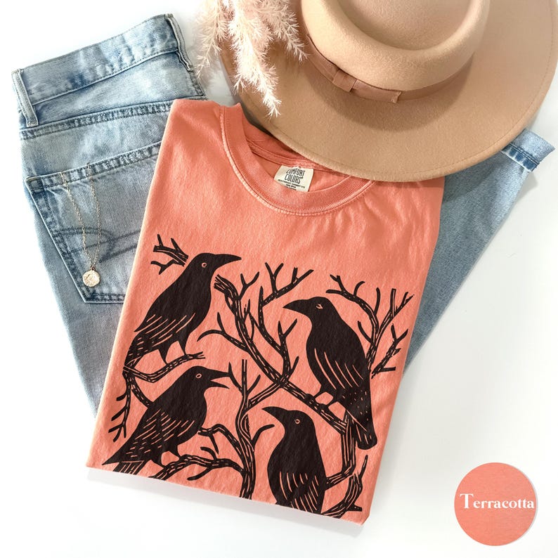 May include: Terracotta-colored t-shirt with a black raven graphic. The shirt is paired with light blue jeans, a tan hat, and a gold necklace. The graphic features four ravens perched on tree branches.
