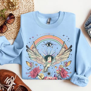 May include: Light blue sweatshirt with a colorful graphic print of a bird with outstretched wings, a rainbow, and flowers. The bird has a large eye and is surrounded by stars and crystals.