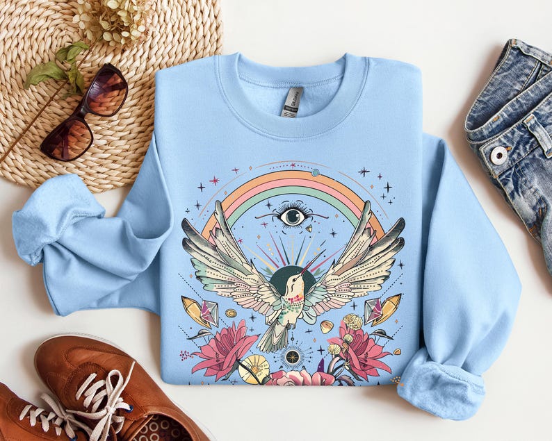 May include: Light blue sweatshirt with a colorful graphic print of a bird with outstretched wings, a rainbow, and flowers. The bird has a large eye and is surrounded by stars and crystals.