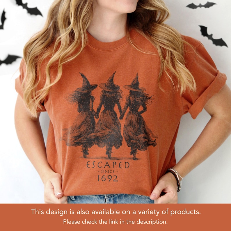 May include: Burnt orange t-shirt featuring a black graphic of three witches holding hands, wearing pointed hats and long dresses. The text "ESCAPED SINCE 1692" is printed below the illustration. The shirt is a comfortable, casual style.