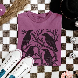 May include: Berry-colored t-shirt featuring a black graphic of four crows perched on tree branches. The shirt is displayed with white sneakers, a black hat, and disco ball ornaments on a checkered surface.