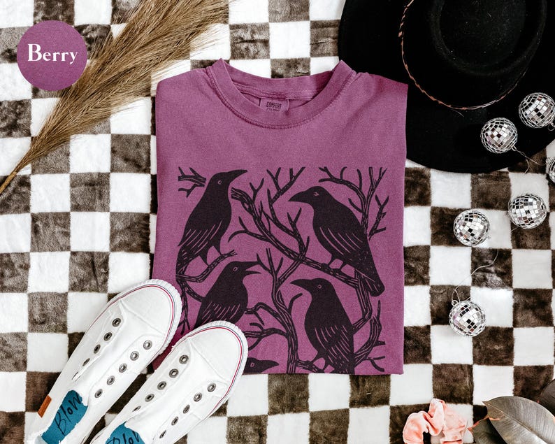 May include: Berry-colored t-shirt featuring a black graphic of four crows perched on tree branches. The shirt is displayed with white sneakers, a black hat, and disco ball ornaments on a checkered surface.