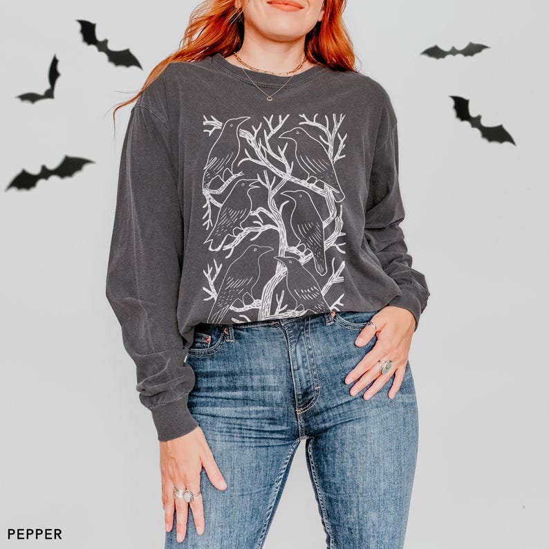 May include: Dark gray long-sleeve shirt featuring a white graphic of crows perched on tree branches. The shirt is paired with blue jeans. The word "PEPPER" is printed in the lower left corner. Black bat cutouts are in the background.