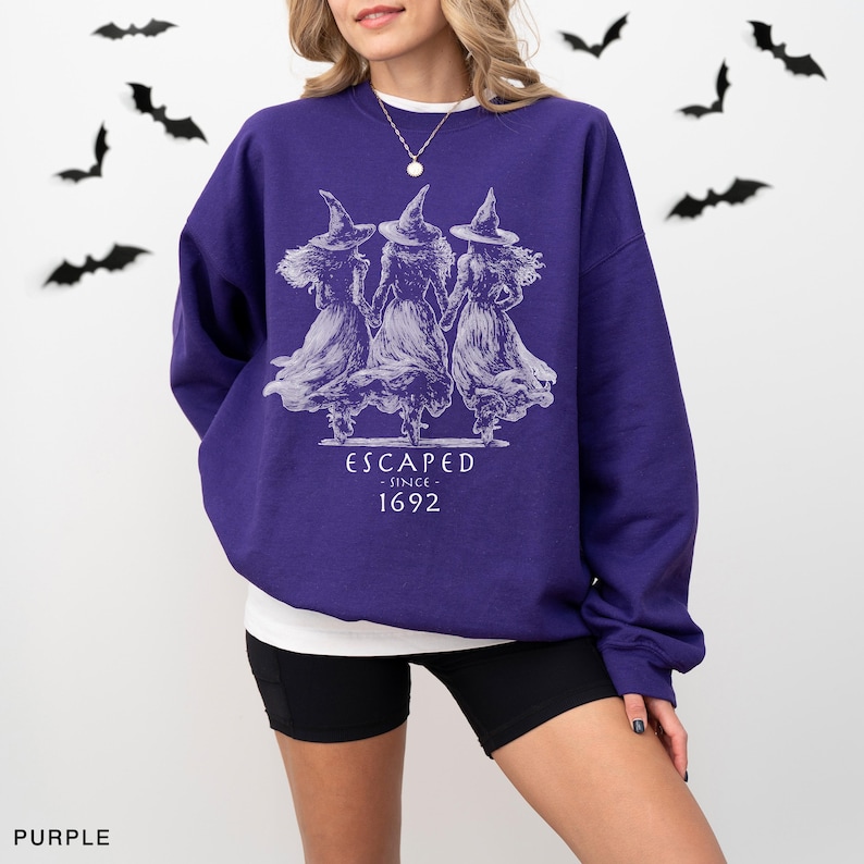 May include: A purple long-sleeve sweatshirt featuring a white graphic of three figures wearing pointed hats and long dresses. The text below the figures reads ESCAPED SINCE 1692. The sweatshirt has a crew neckline and a relaxed fit.