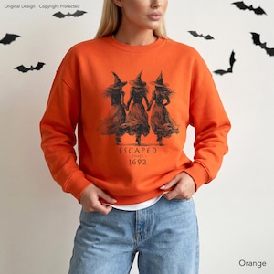 May include: An orange long-sleeve sweatshirt featuring a black graphic print of three witches holding hands. The text below the figures reads ESCAPED SINCE 1692. The garment has a crew neckline and ribbed cuffs, paired with blue denim jeans.