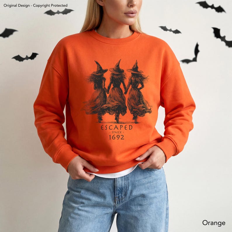 May include: An orange long-sleeve sweatshirt featuring a black graphic print of three witches holding hands. The text below the figures reads ESCAPED SINCE 1692. The garment has a crew neckline and ribbed cuffs, paired with blue denim jeans.