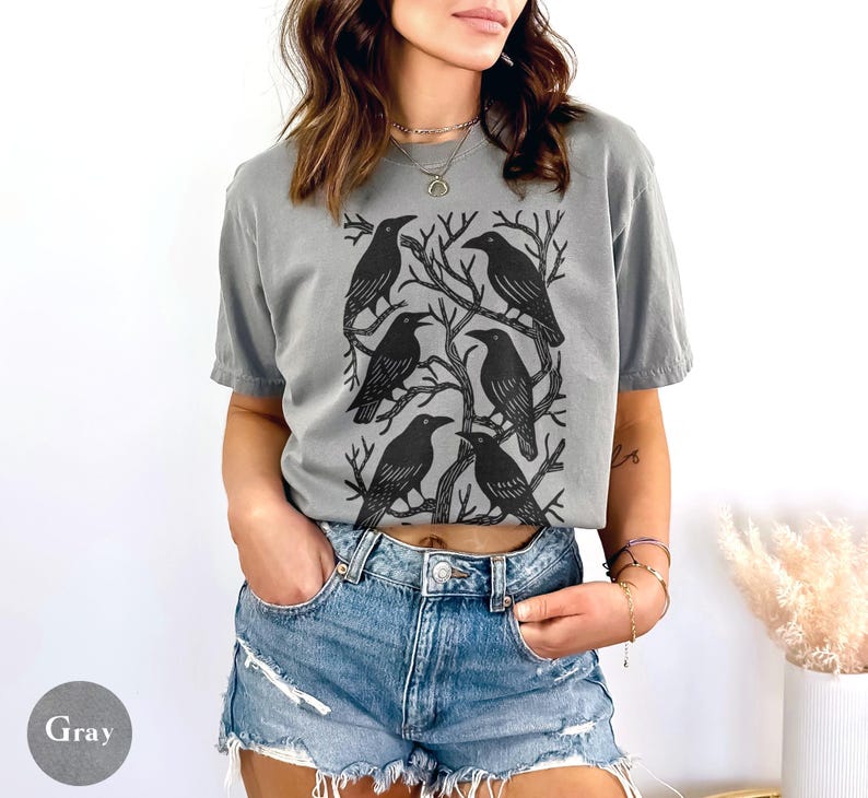 May include: Gray t-shirt featuring a black graphic of six crows perched on tree branches. The shirt is a relaxed fit and has short sleeves. The word "Gray" is printed on a gray circle in the lower left corner.