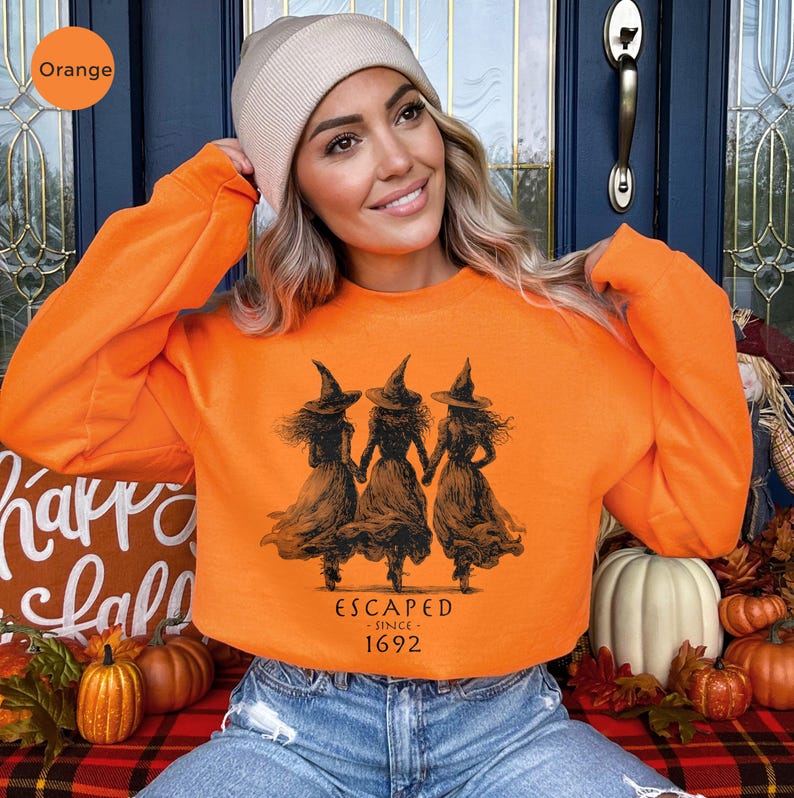 May include: An orange long-sleeve sweatshirt featuring a black graphic of three witches wearing pointed hats and long dresses, holding hands. Below the illustration, the text reads "ESCAPED - SINCE - 1692" in a simple black font.