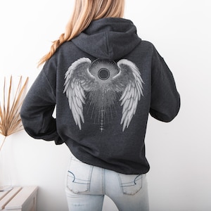 May include: Dark gray hoodie with a large white angel wing design on the back. The graphic includes detailed feathering, a central sunburst, and a series of vertical lines. The hoodie has a drawstring hood and a kangaroo pocket.