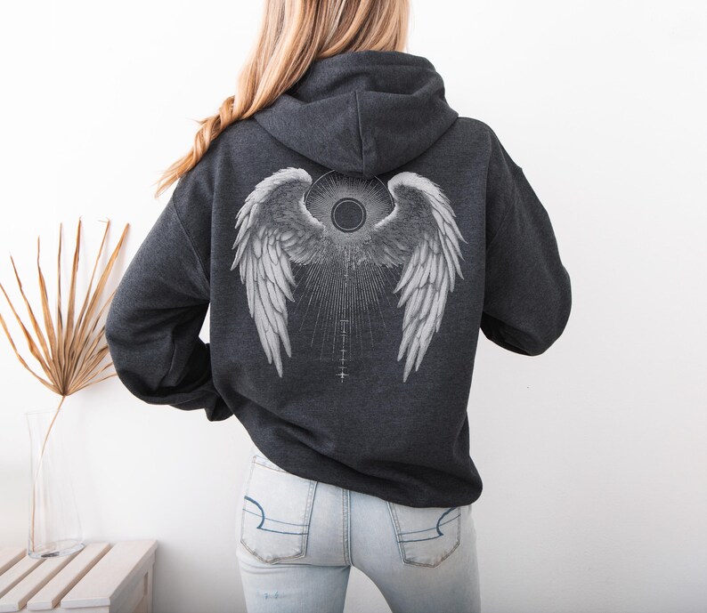 May include: Dark gray hoodie with a large white angel wing design on the back. The graphic includes detailed feathering, a central sunburst, and a series of vertical lines. The hoodie has a drawstring hood and a kangaroo pocket.