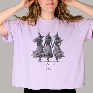 May include: A light purple t-shirt featuring a graphic of three witches holding hands, wearing pointed hats and flowing dresses. The text "ESCAPED since 1692" is printed below the graphic. The shirt is a cropped style.