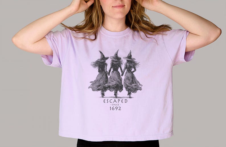 May include: A light purple t-shirt featuring a graphic of three witches holding hands, wearing pointed hats and flowing dresses. The text "ESCAPED since 1692" is printed below the graphic. The shirt is a cropped style.