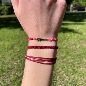May include: A close-up of a person's wrist wearing three bracelets. The top bracelet is a silver charm bracelet with the word "Kismet" on it. The middle bracelet is a dark red braided cord bracelet. The bottom bracelet is a thin, dark red cord bracelet with a white cord woven through it.