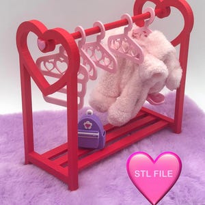 Puede incluir: Un perchero rojo para muñecas con soportes laterales en forma de corazón y un estante. Perchas rosas sostienen un conjunto rosa esponjoso. Una pequeña mochila morada está en el estante. La imagen incluye un corazón rosa con el texto "STL FILE".