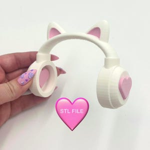 May include: White headphones with pink accents, featuring cat ears and heart-shaped details. The ear cups and the heart-shaped accents are pink. The text "STL FILE" is on a pink heart.