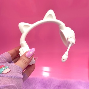 May include: White cat ear headphones with a flexible microphone. The headphones have a headband with two cat ears on top and ear cups. The microphone is attached to the right ear cup. The background is a solid pink color.