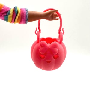 May include: A bright pink pumpkin-shaped basket with a handle, bow, and heart-shaped cutouts. The basket is held by a person wearing a colorful striped sleeve. The basket is likely for Halloween or a similar holiday.