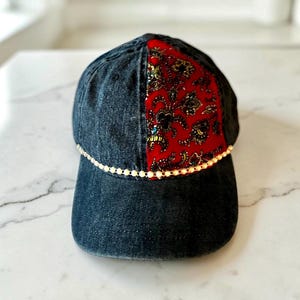 May include: A dark charcoal denim baseball cap featuring a central vertical panel of red fabric with a detailed black and gold floral pattern. A thin, delicate string of small, light-colored beads runs horizontally across the front of the cap.