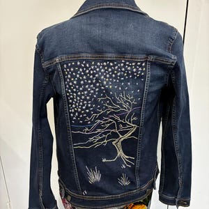 May include: A dark blue denim jacket featuring an embroidered design on the back. The embroidery depicts a bare tree with sprawling branches beneath a night sky filled with numerous small white and yellow stars, with small tufts of grass at the base.