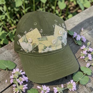 May include: An olive green baseball cap with a floral design. The cap features a patch of yellow fabric with white floral embroidery and two beige buttons. The cap is sitting on a wooden surface with purple flowers.