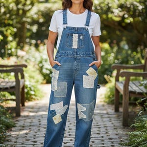 May include: Blue denim overalls featuring a white crew neck t-shirt underneath. The overalls have a medium wash, metal hardware, and multiple decorative patches made of white and cream lace and floral fabric on the bib and pant legs.
