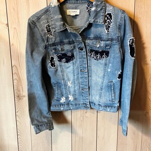 May include: A light blue denim jacket featuring distressed areas and black paisley bandana fabric patches. The jacket has a button-up front, two chest pockets, and a collar. The interior label reads CI SONO. The garment is displayed on a wooden hanger.