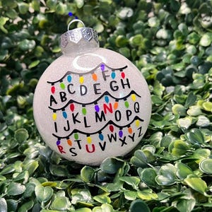May include: A round, white glittery Christmas ornament featuring a black string of holiday lights draped in three rows. The alphabet is printed in black letters beneath the lights, with some letters highlighted in red, blue, orange, and green.