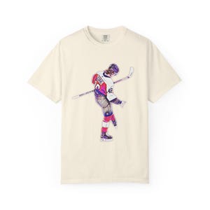 May include: Cream-colored t-shirt featuring a watercolor illustration of a hockey player in action. The player wears a red, white, and purple uniform with the number 46. The hockey stick is held in the player's hands.