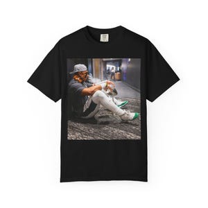 May include: Black t-shirt featuring a photograph of a person sitting on the ground, exhaling smoke. The person is wearing a gray cap, gray shirt, white pants with a dark stripe, and white sneakers with green accents.