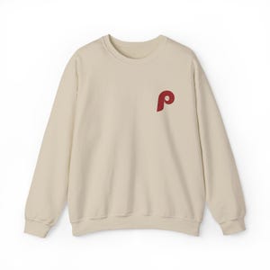 May include: A cream-colored sweatshirt with a red embroidered "P" logo on the upper left chest. The long-sleeved crew neck top is made of a soft material and is suitable for casual wear.