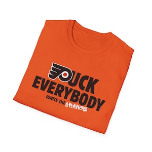 Può includere: T-shirt arancione con la scritta "DUCK EVERYBODY" in nero e "IGNITE THE ORANGE" in bianco. La maglietta presenta anche un logo con un'ala e un cerchio. La maglietta è a girocollo.