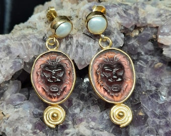 May include: A pair of gold earrings with a pink cameo featuring a lion's face and a white pearl accent.